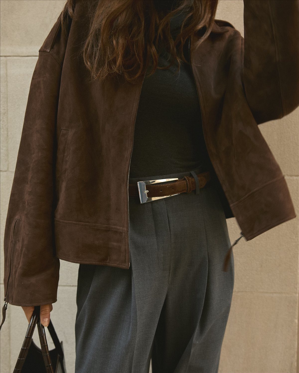 A person wearing a brown suede jacket, black turtleneck, and grey trousers with a silver buckle belt.