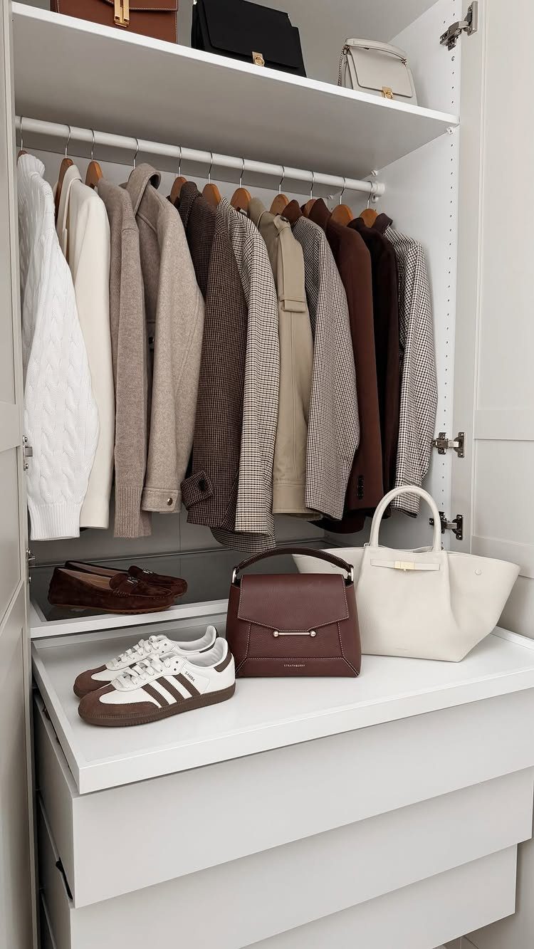 Neutral colored clothing hanging in a white closet with handbags and sneakers on the shelf below.