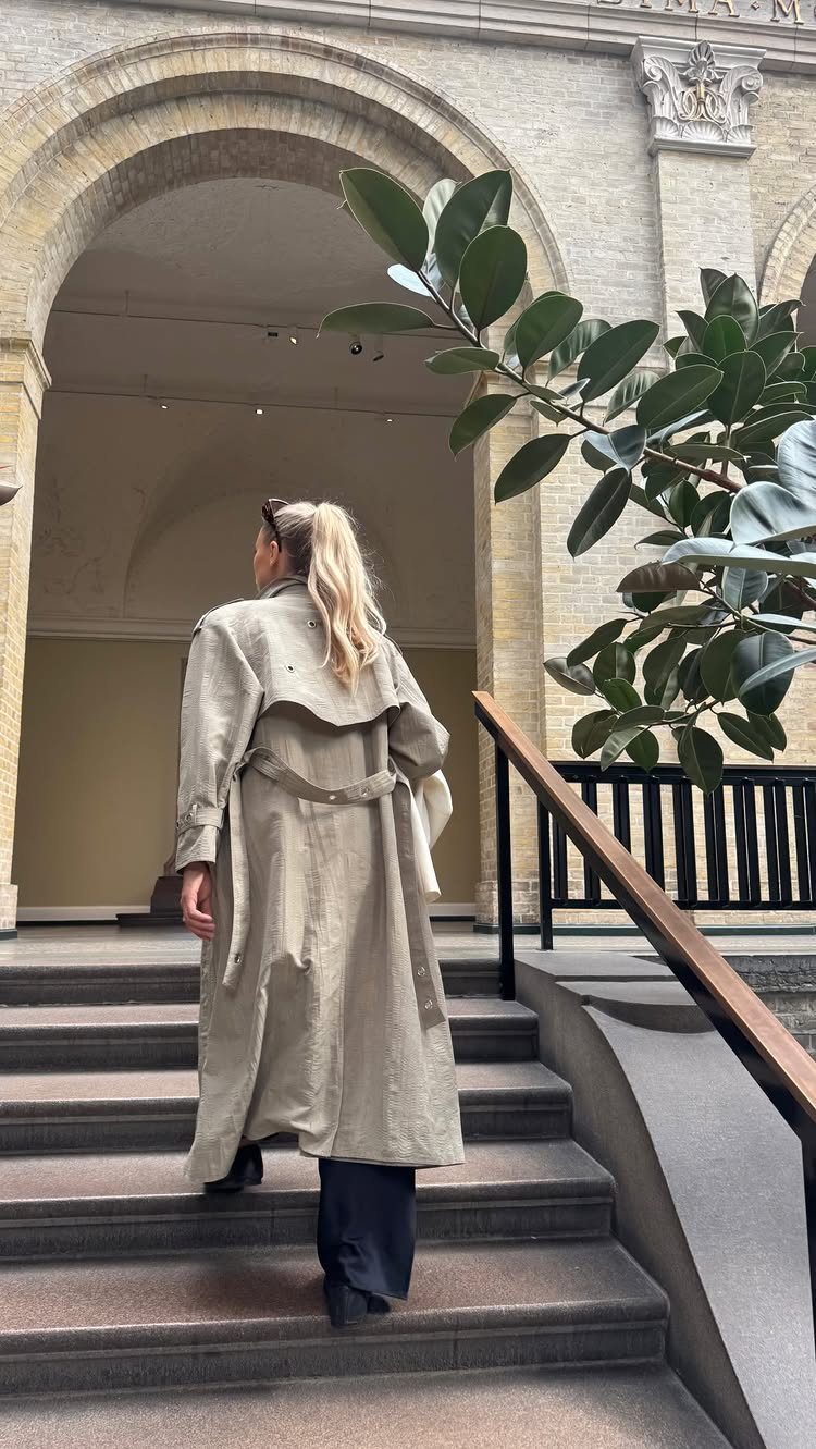 A woman wearing a long beige trench coat walking up stone stairs towards an archway.
