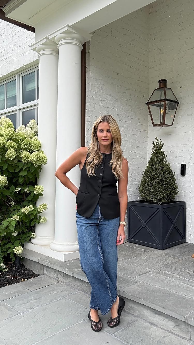 A woman posing in a black vest and blue jeans in front of a white house.