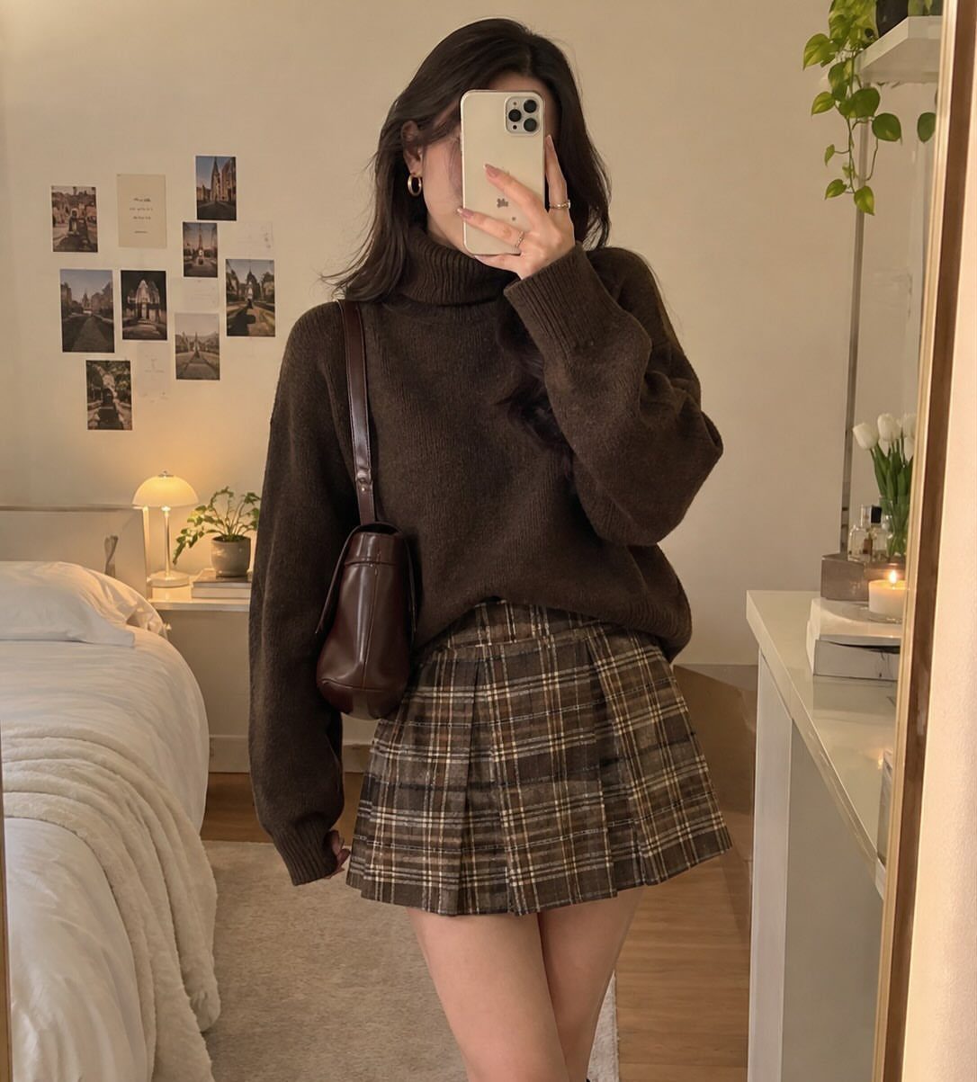 A person wearing a brown turtleneck sweater and a plaid pleated skirt posing for a mirror selfie.