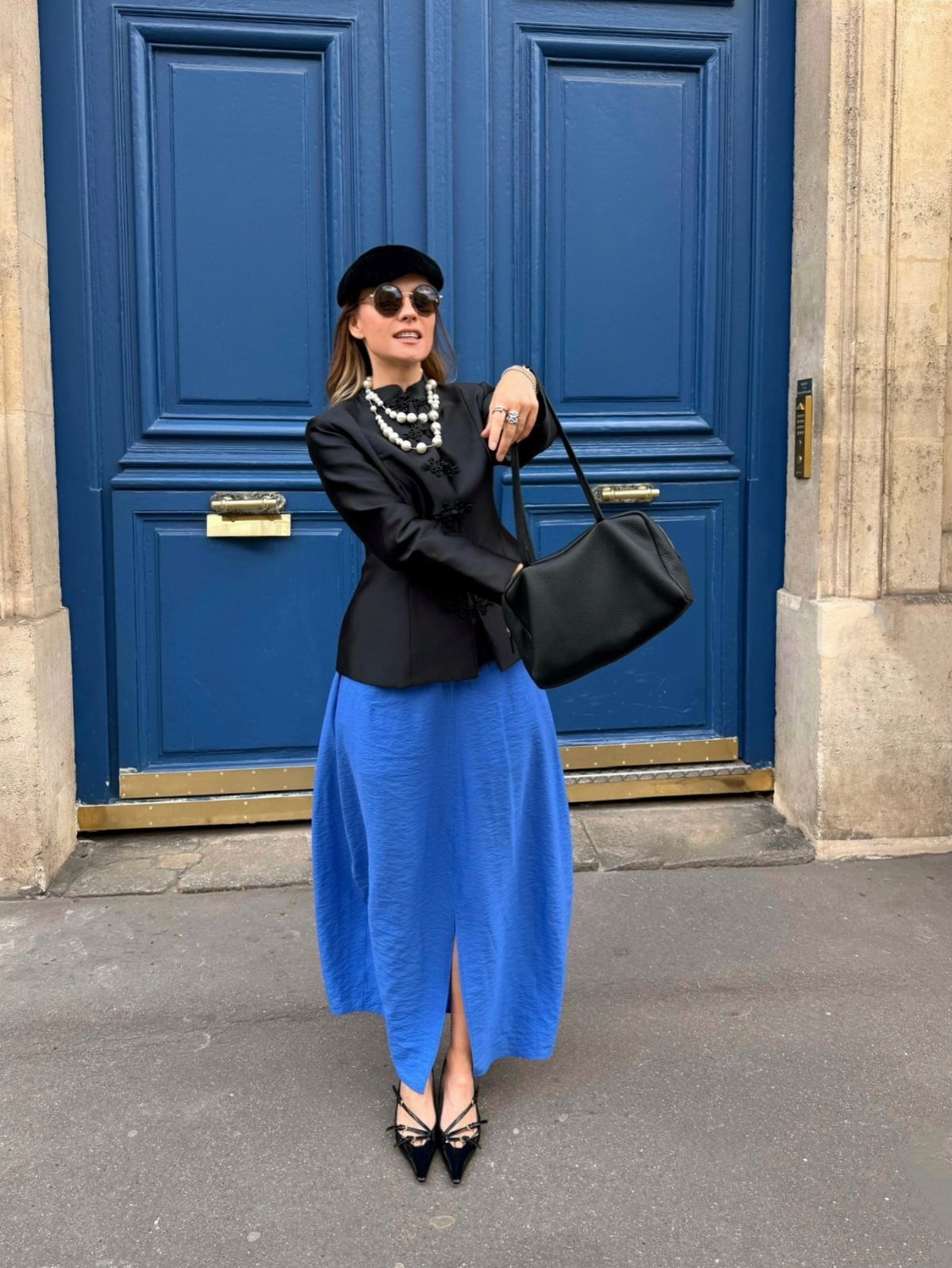 A woman wearing a black blazer and long blue skirt poses in front of blue doors.