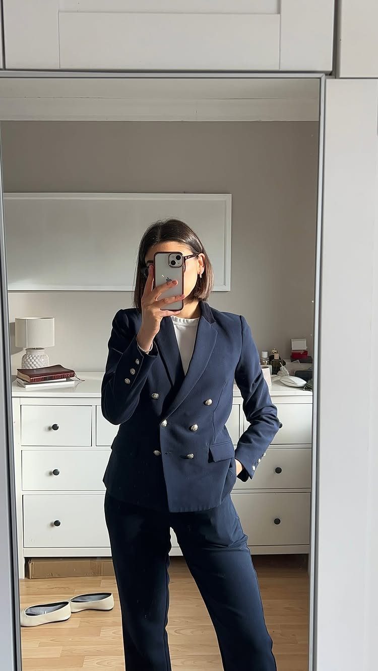 Woman in a navy blue double-breasted blazer and matching trousers taking a mirror selfie.