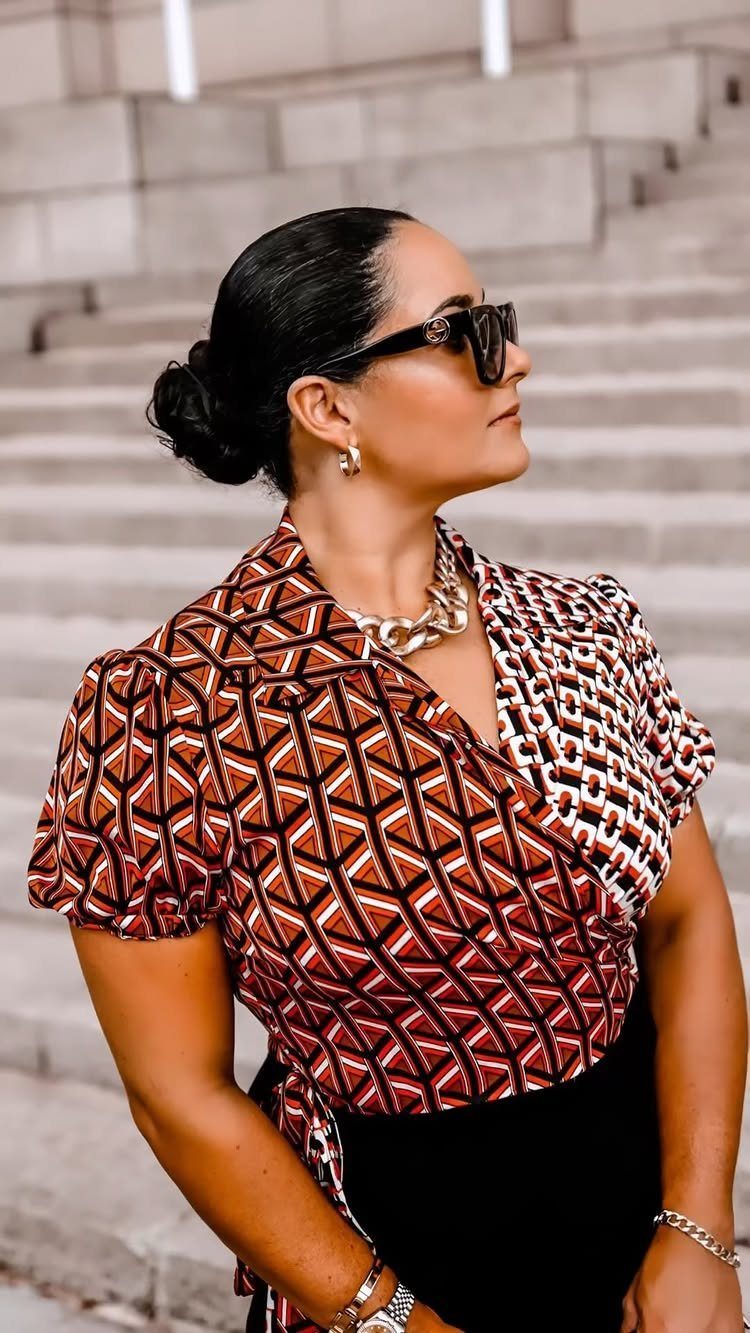 A woman in a patterned geometric wrap blouse and sunglasses posing outdoors.
