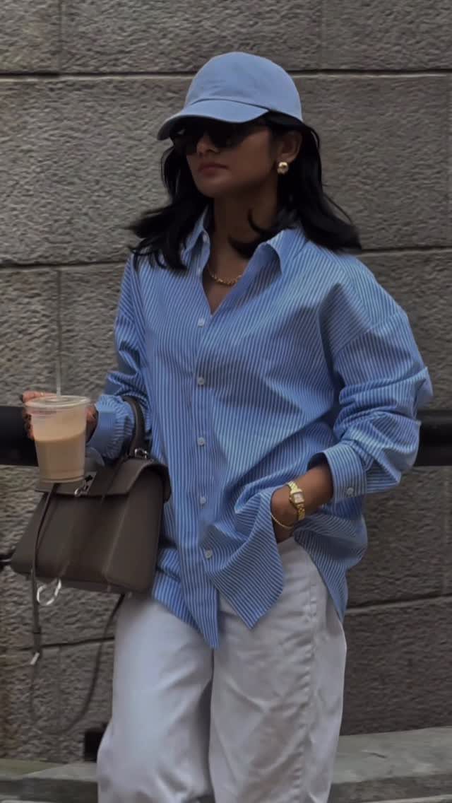 Woman in a blue striped shirt, white pants, and a light blue cap holding coffee.