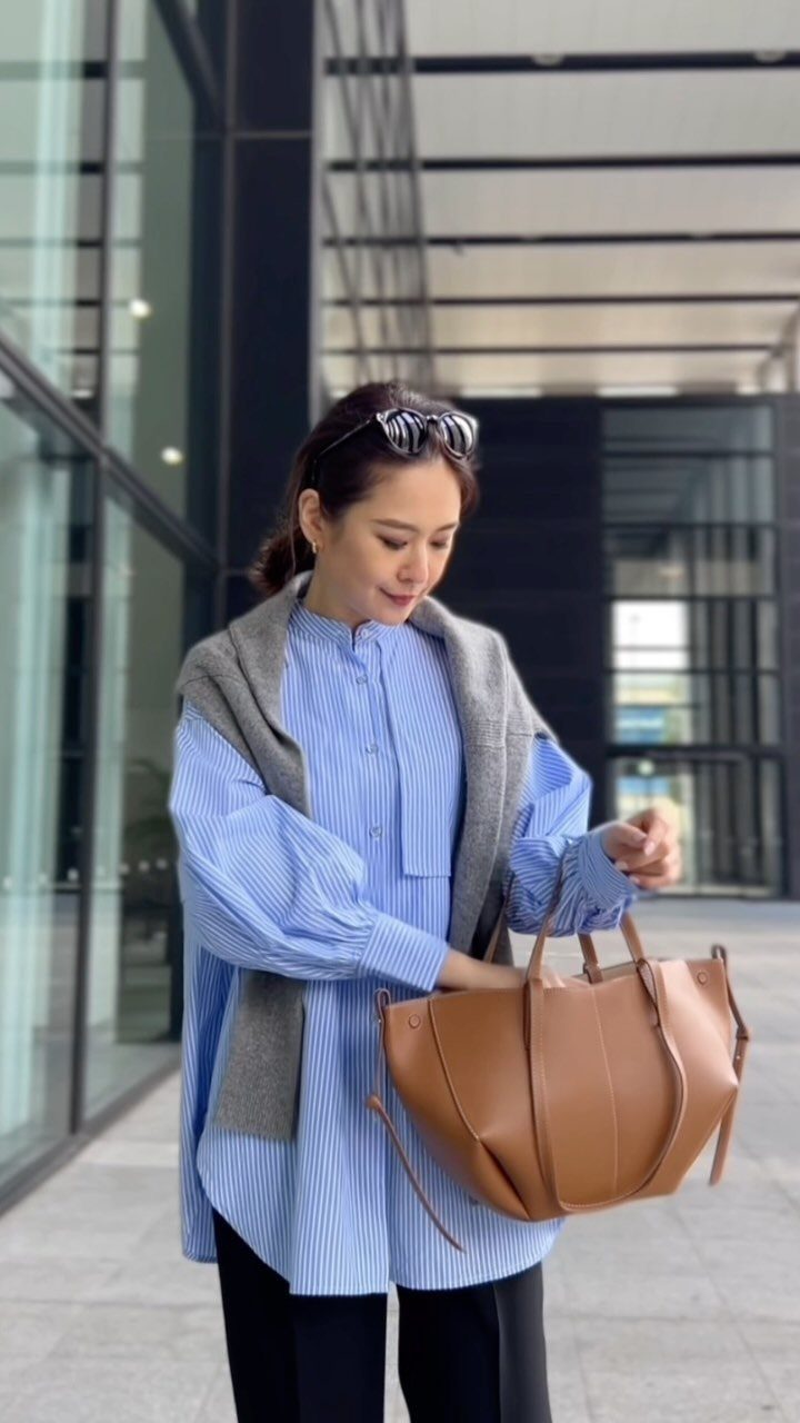 Woman in a blue striped shirt, grey vest, and black trousers holding a tan handbag.