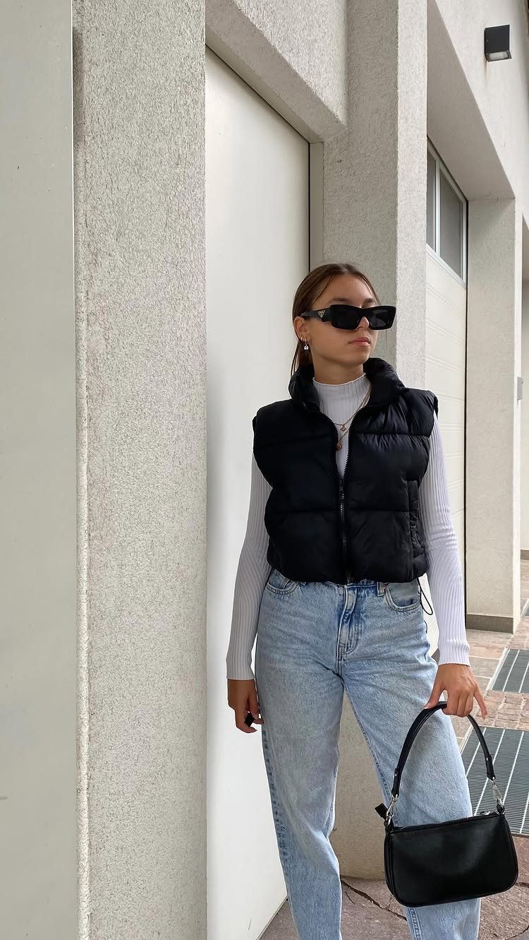 Woman in a black puffer vest, white top, and light blue jeans holding a black handbag.
