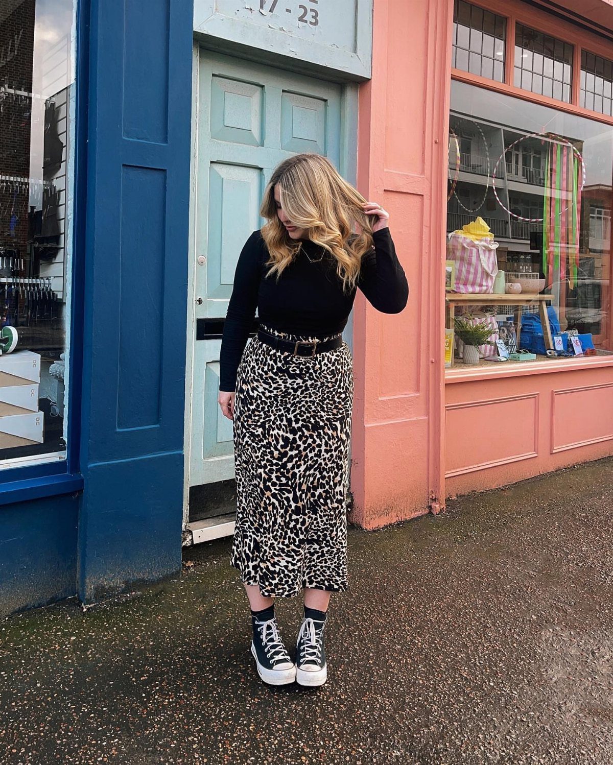 Woman wearing a black fitted top and leopard print midi skirt with black sneakers.