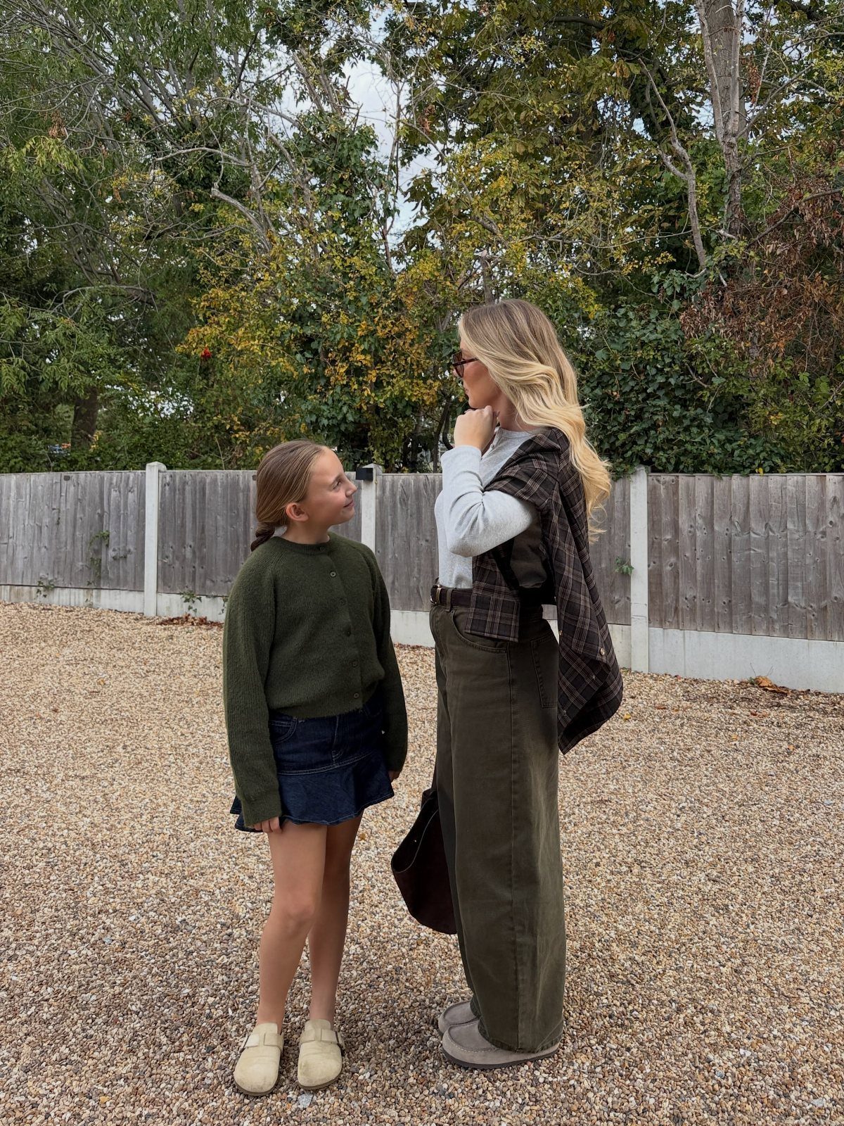 A woman and a young girl standing together outdoors wearing casual olive and denim clothing.