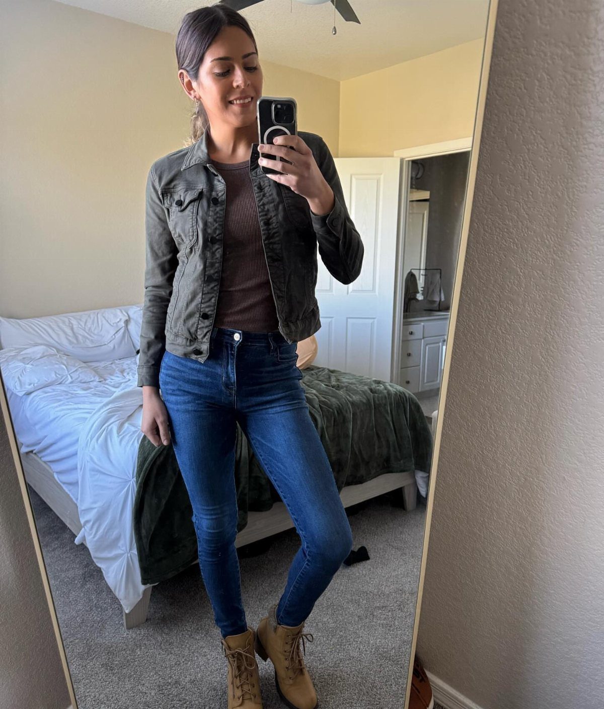 Woman in olive jacket, brown top, blue jeans, and tan boots posing for a mirror selfie.