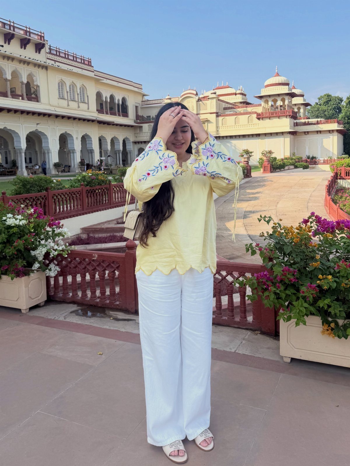 Woman in a yellow floral embroidered blouse and white wide-leg trousers standing in a palace courtyard.