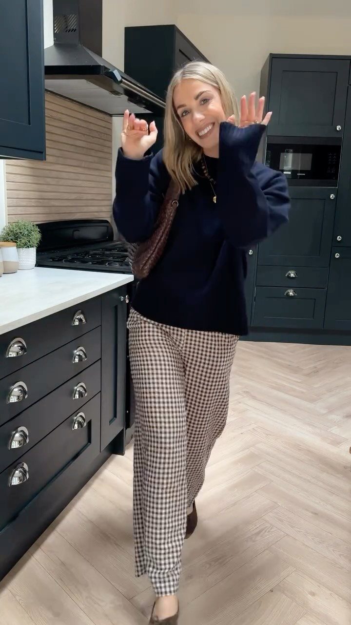 A smiling woman wearing a navy sweater and checkered patterned tailored trousers in a kitchen.