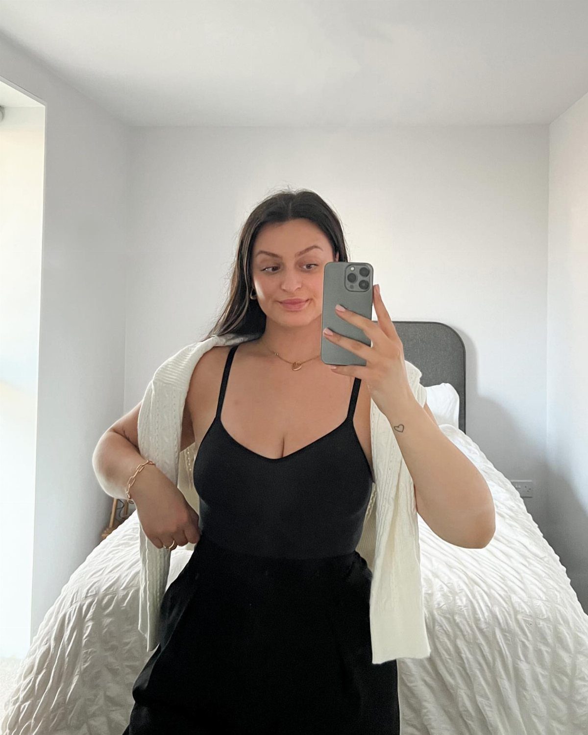 Woman in a black sleeveless bodysuit and white cardigan taking a mirror selfie in a bedroom.