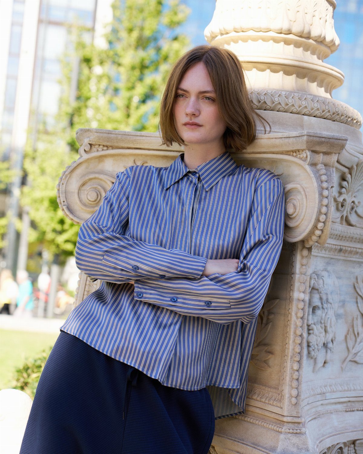 A woman wearing a blue and white striped button-down shirt leaning against a stone pillar.