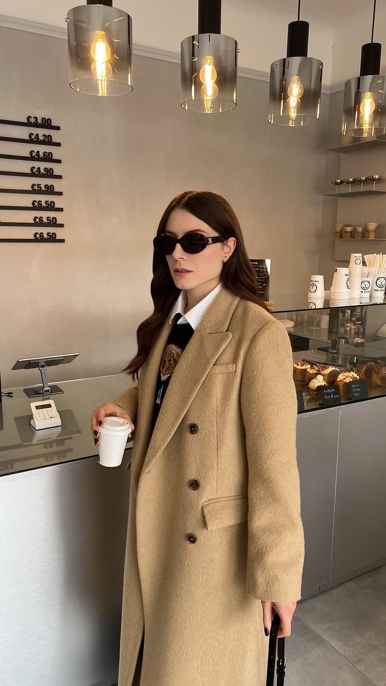 A woman in sunglasses and a long camel coat holding a coffee cup in a cafe.