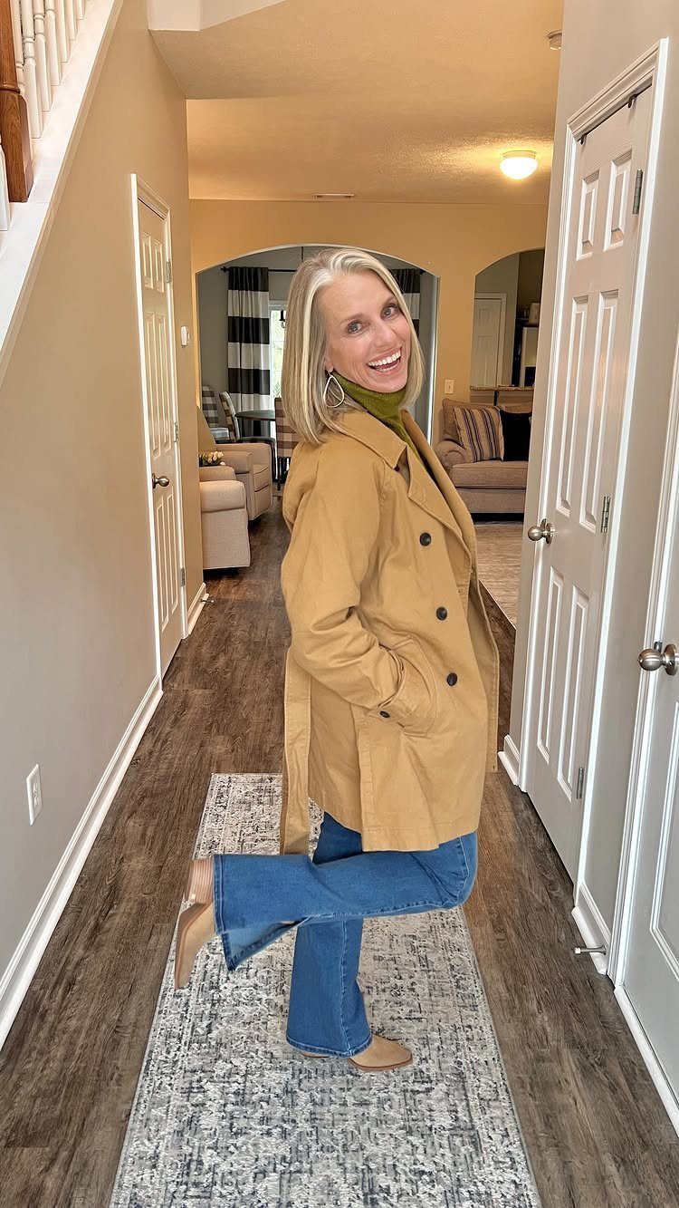 Smiling blonde woman wearing a tan trench coat, green turtleneck, and blue flared jeans in a hallway.