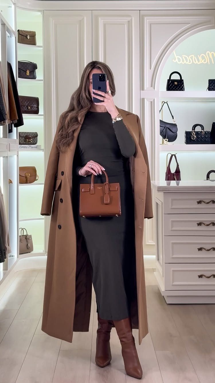 Woman in a long dark green dress, tan trench coat, and brown boots holding a handbag.