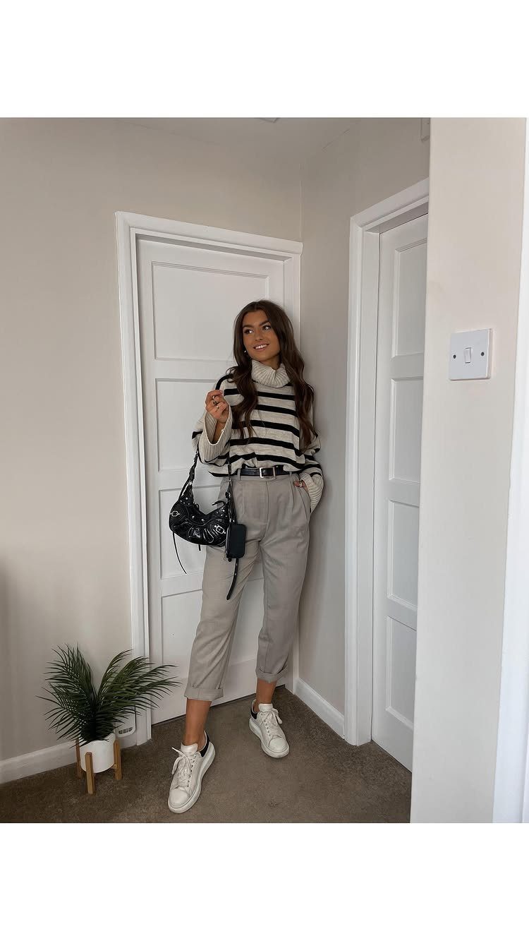 A woman posing in a striped sweater, grey trousers, and white sneakers in a hallway.