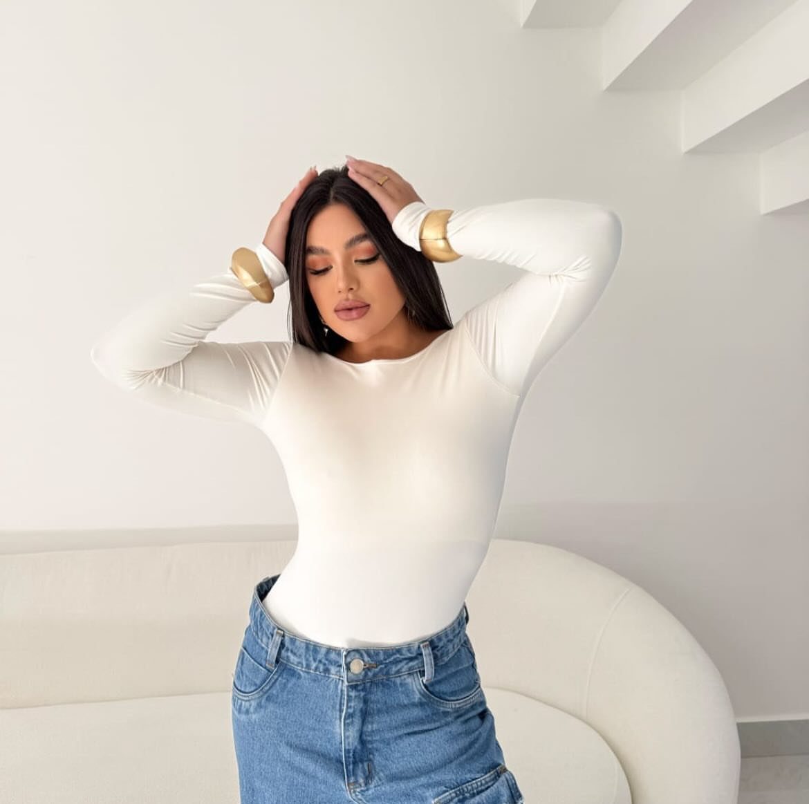 Woman in a white long-sleeved bodysuit and blue jeans posing with hands on her head.