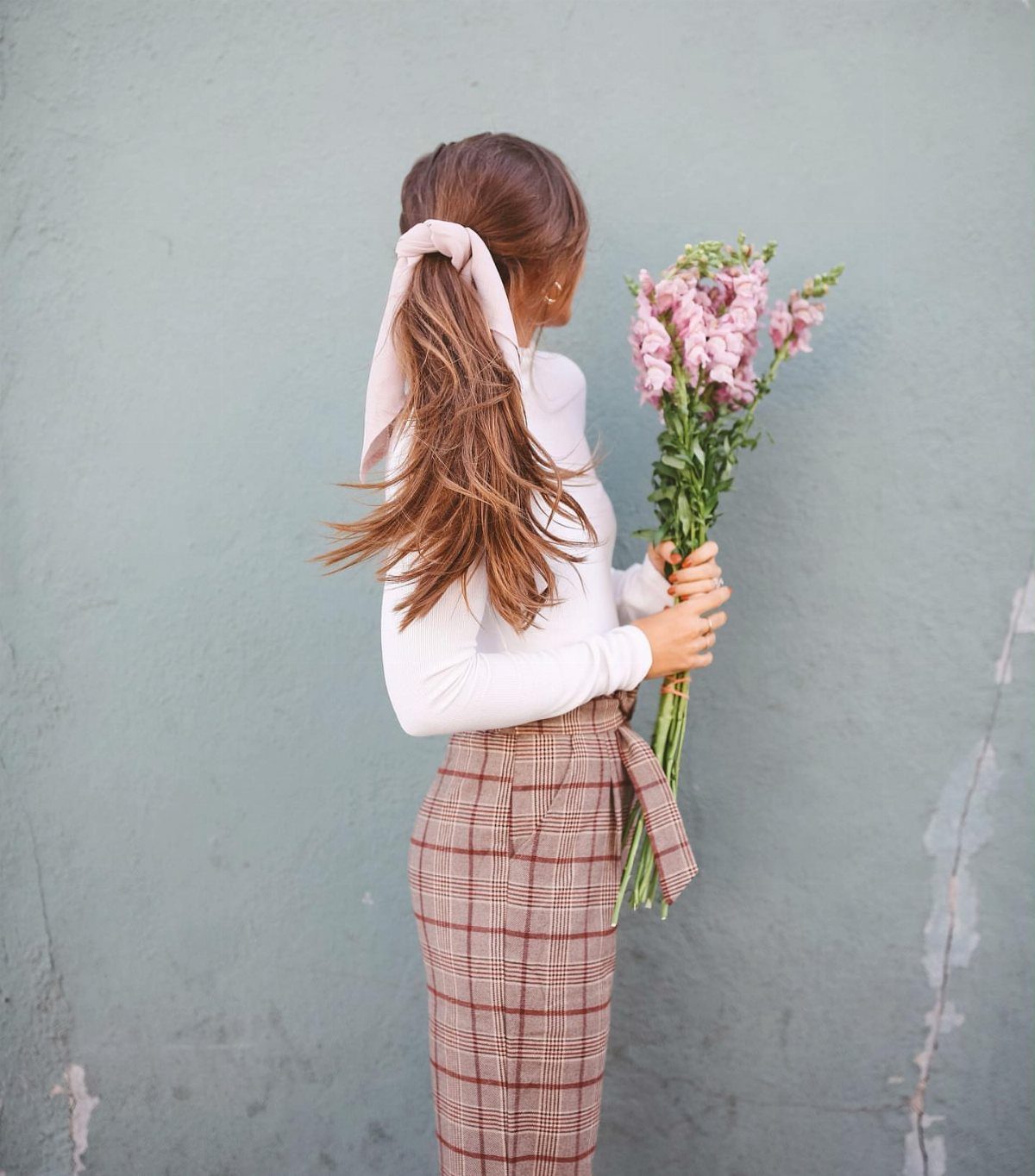 Woman in a white ribbed turtleneck and plaid trousers holding pink flowers against a blue wall.