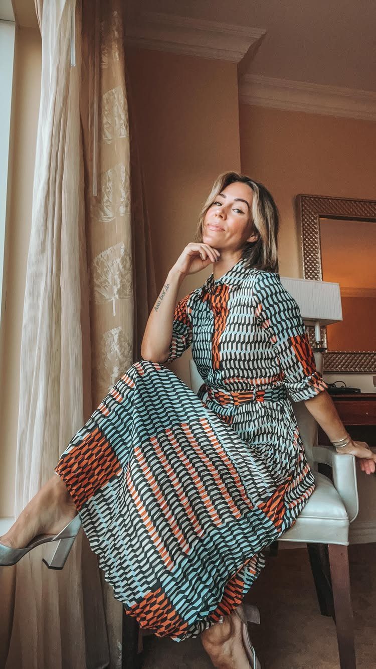 A woman in a patterned teal and orange midi dress sitting by a window.