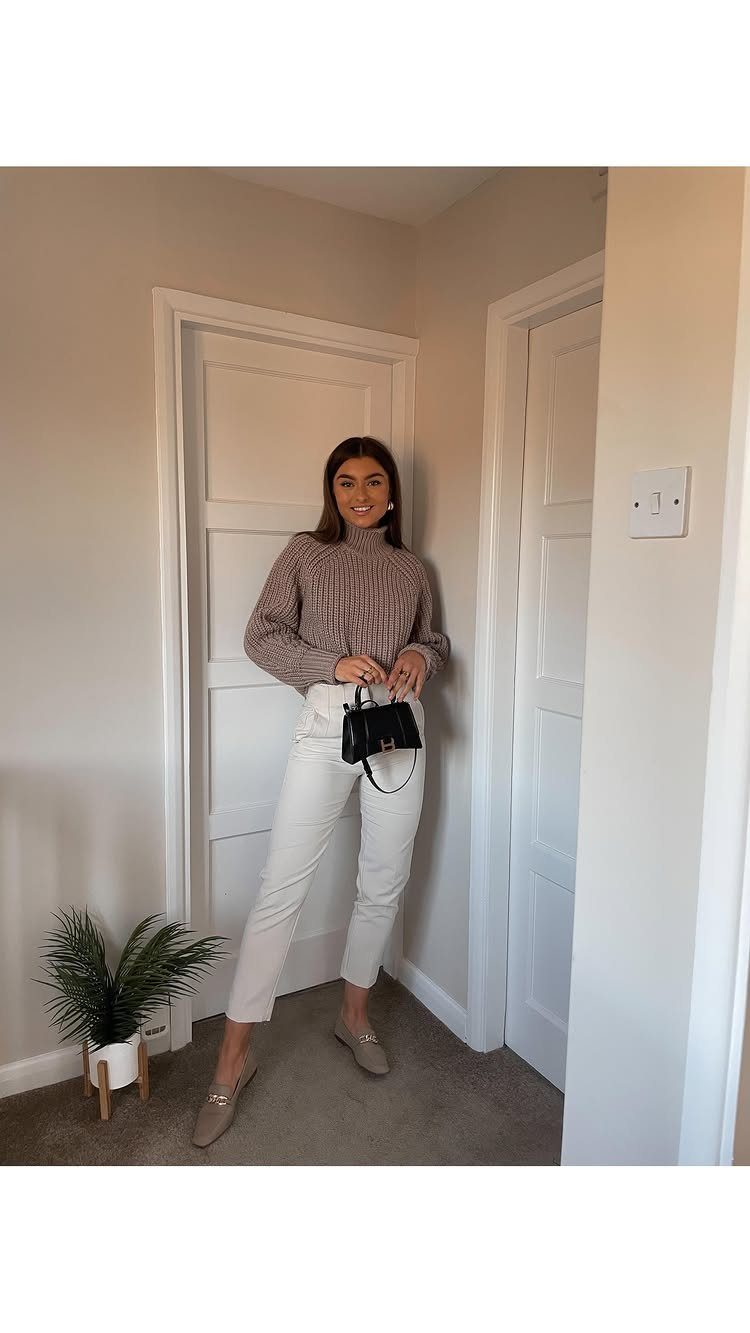 A woman wearing a brown turtleneck, white trousers, and beige flats posing in a hallway.