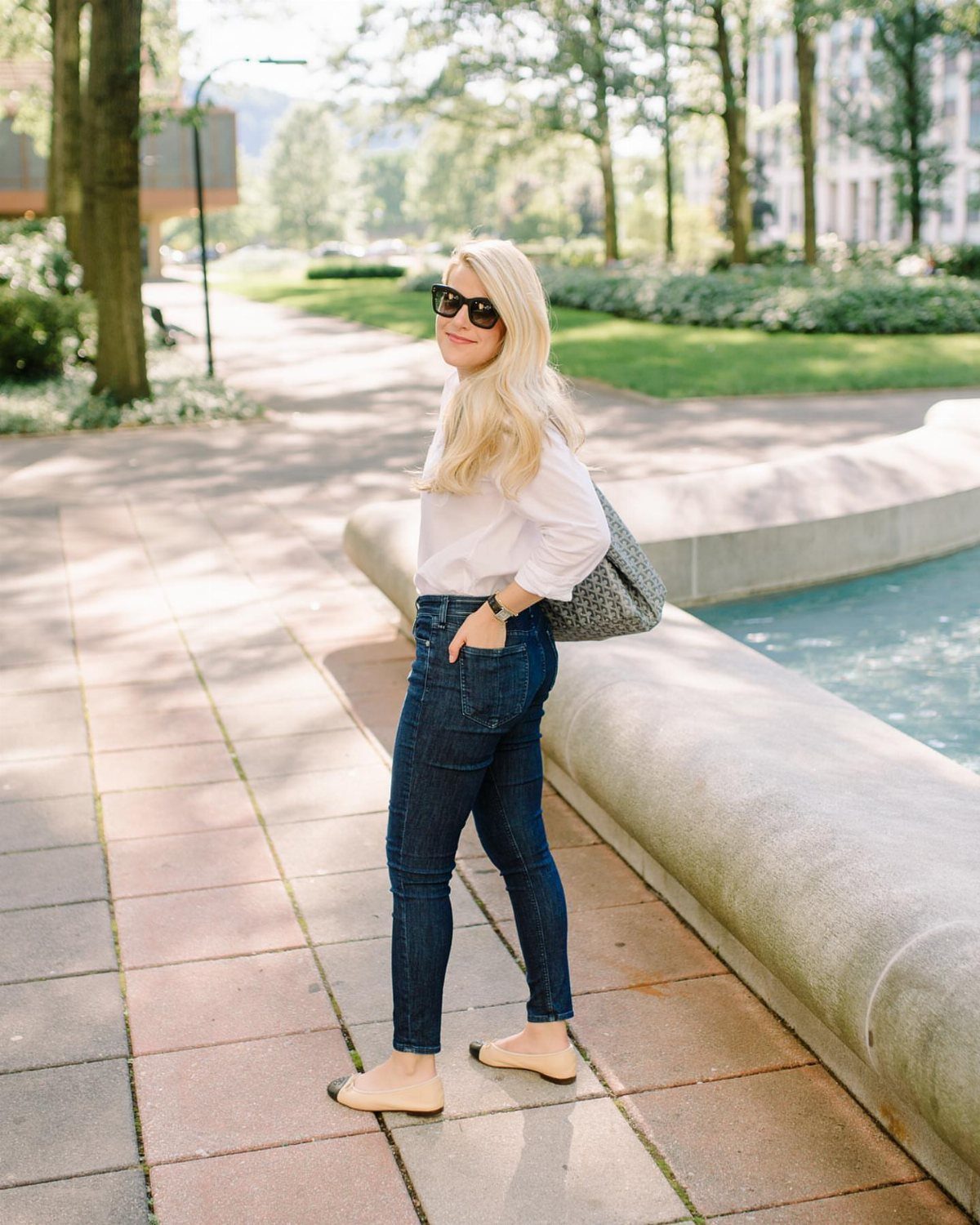 A blonde woman posing in a white blouse and dark wash jeans by a fountain.
