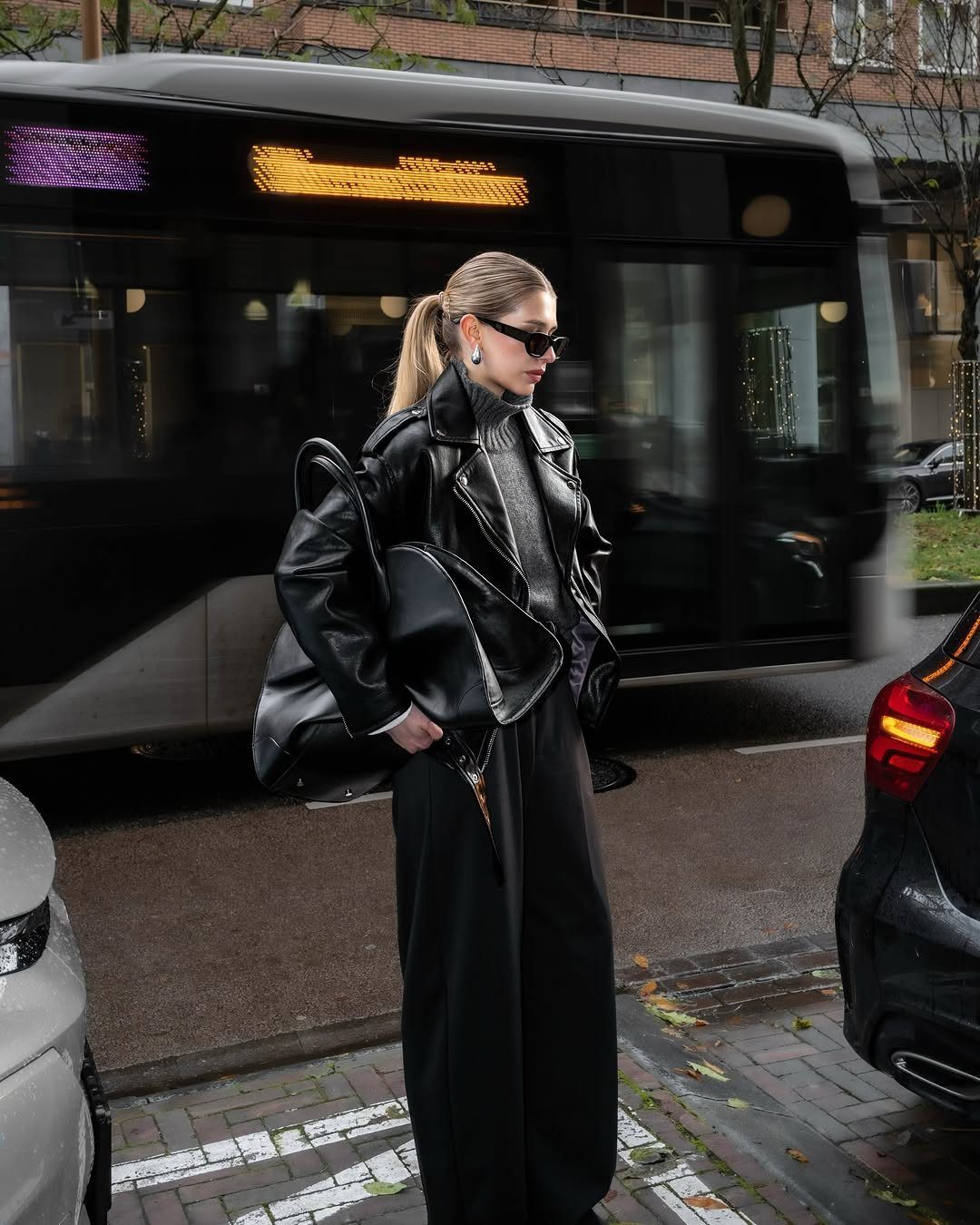 A woman in a black leather jacket and wide-leg trousers walking on a city street.