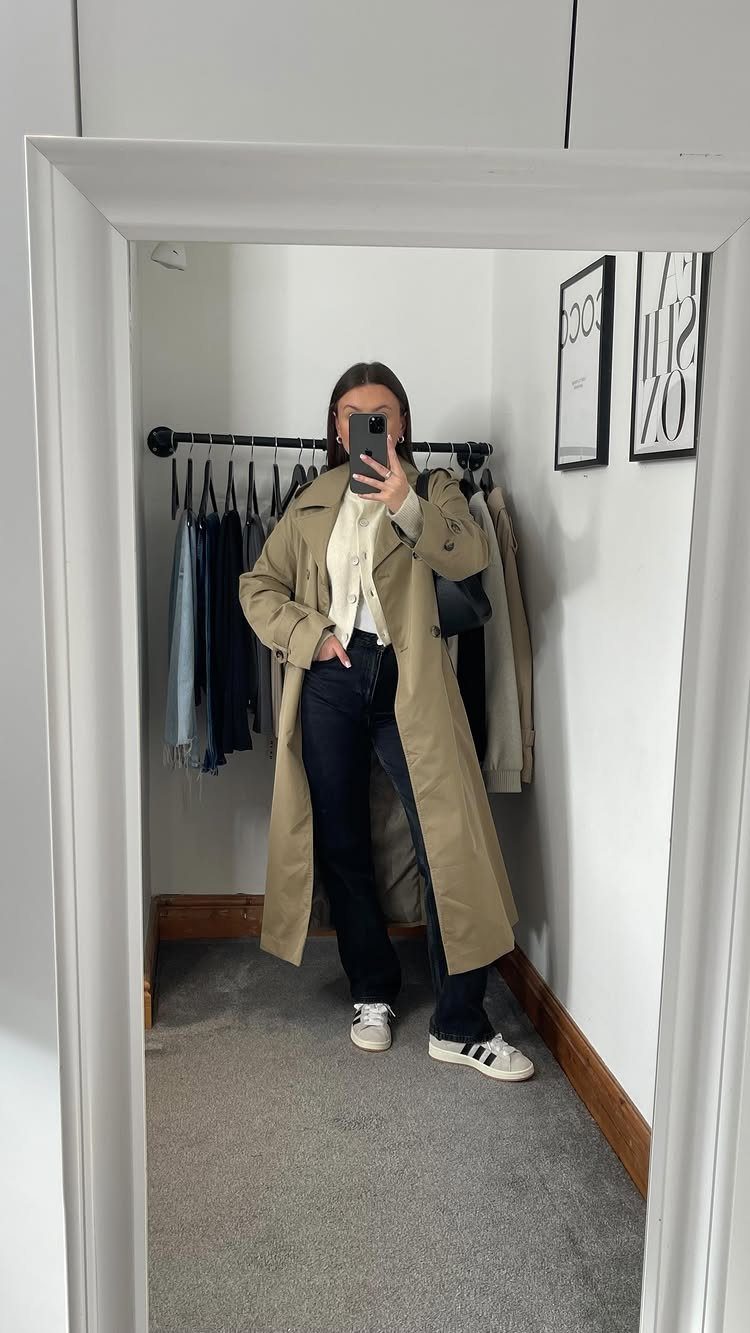 A woman posing in a mirror wearing a long tan trench coat and dark jeans.