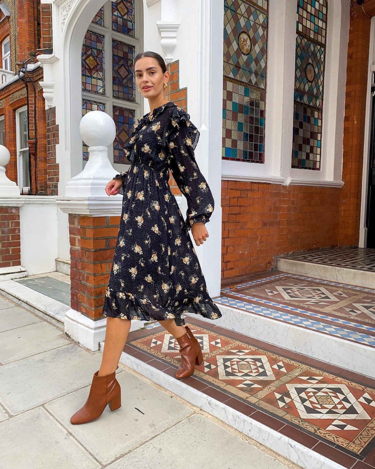 A woman in a black floral dress walking while wearing brown leather ankle boots.