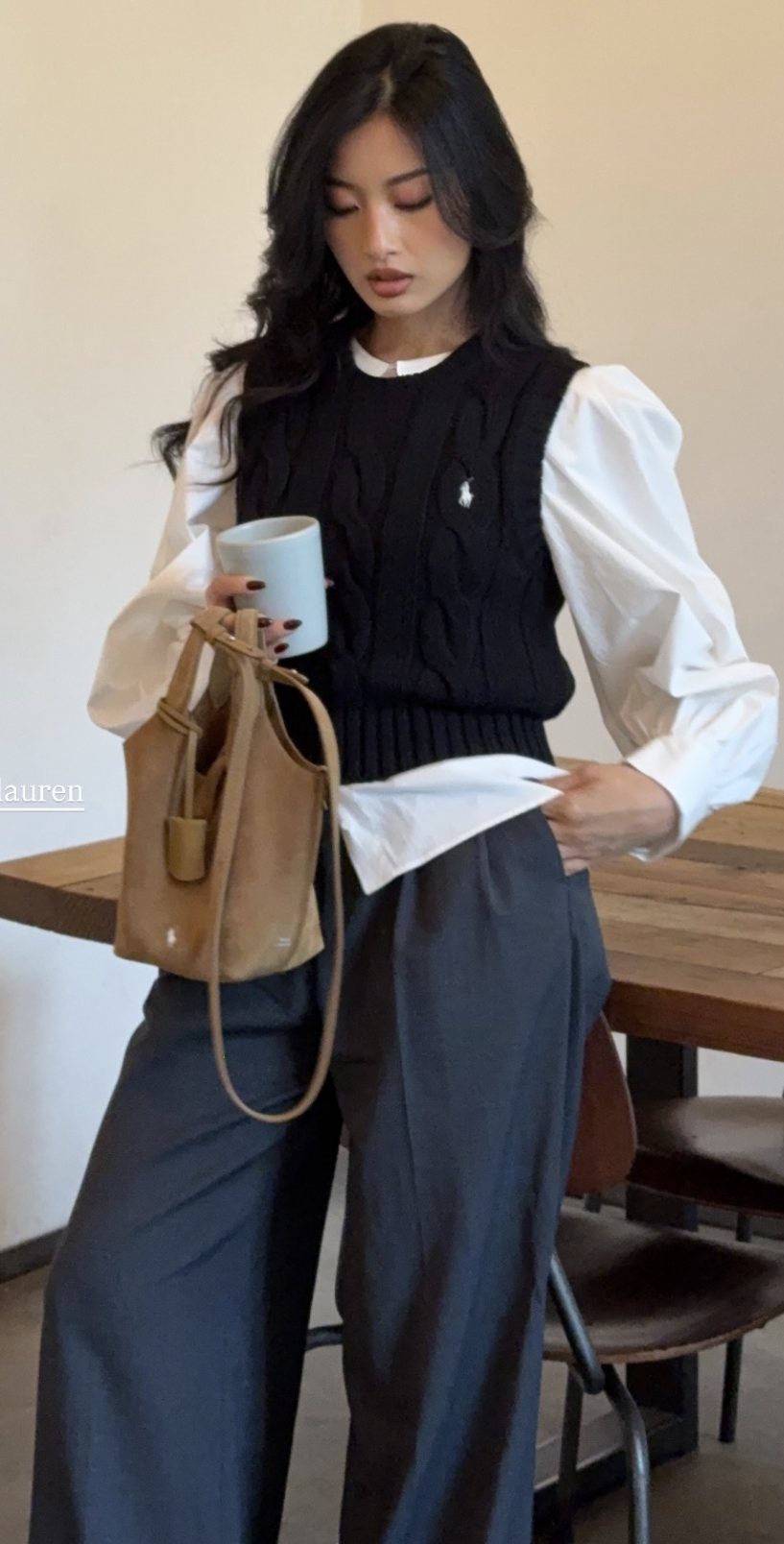 Woman wearing a black cable-knit sweater vest over a white shirt and grey trousers.