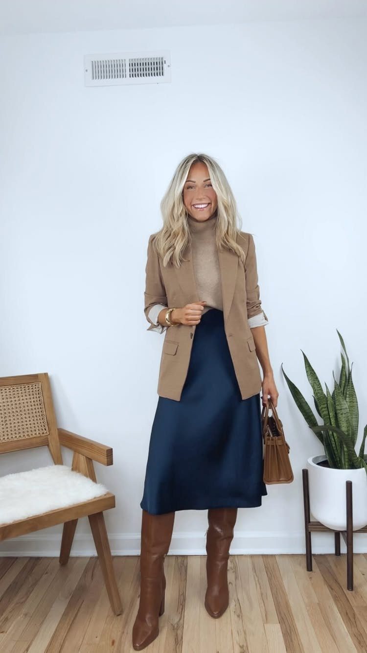 Smiling woman wearing a tan blazer, beige turtleneck, navy satin skirt, and brown leather boots.