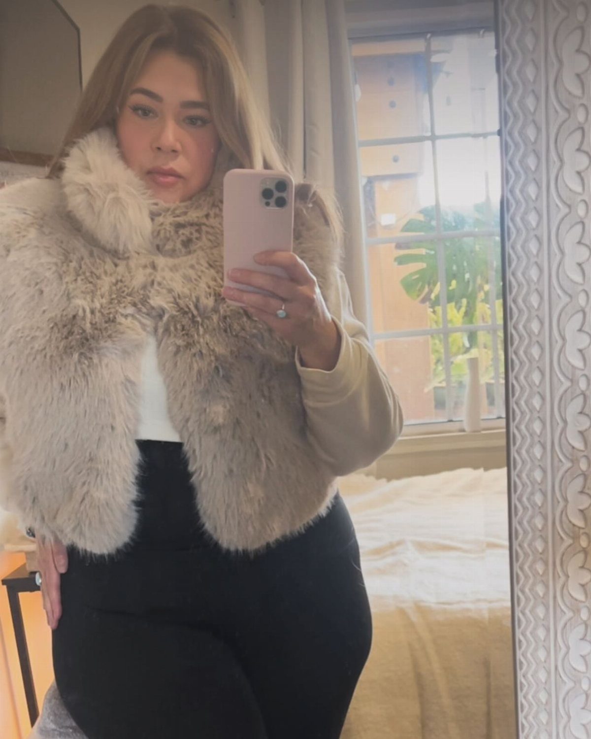 A woman posing in a mirror wearing a light brown furry vest and black pants.