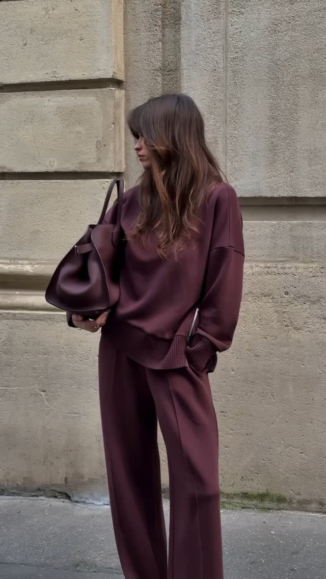A woman in a maroon coordinated knit set carrying a large leather handbag against a stone wall.