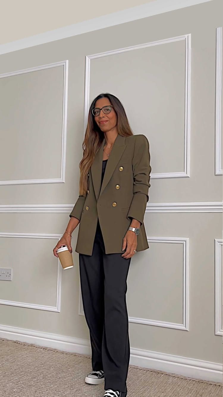 Woman in olive green blazer and black wide-leg trousers standing against a paneled wall.