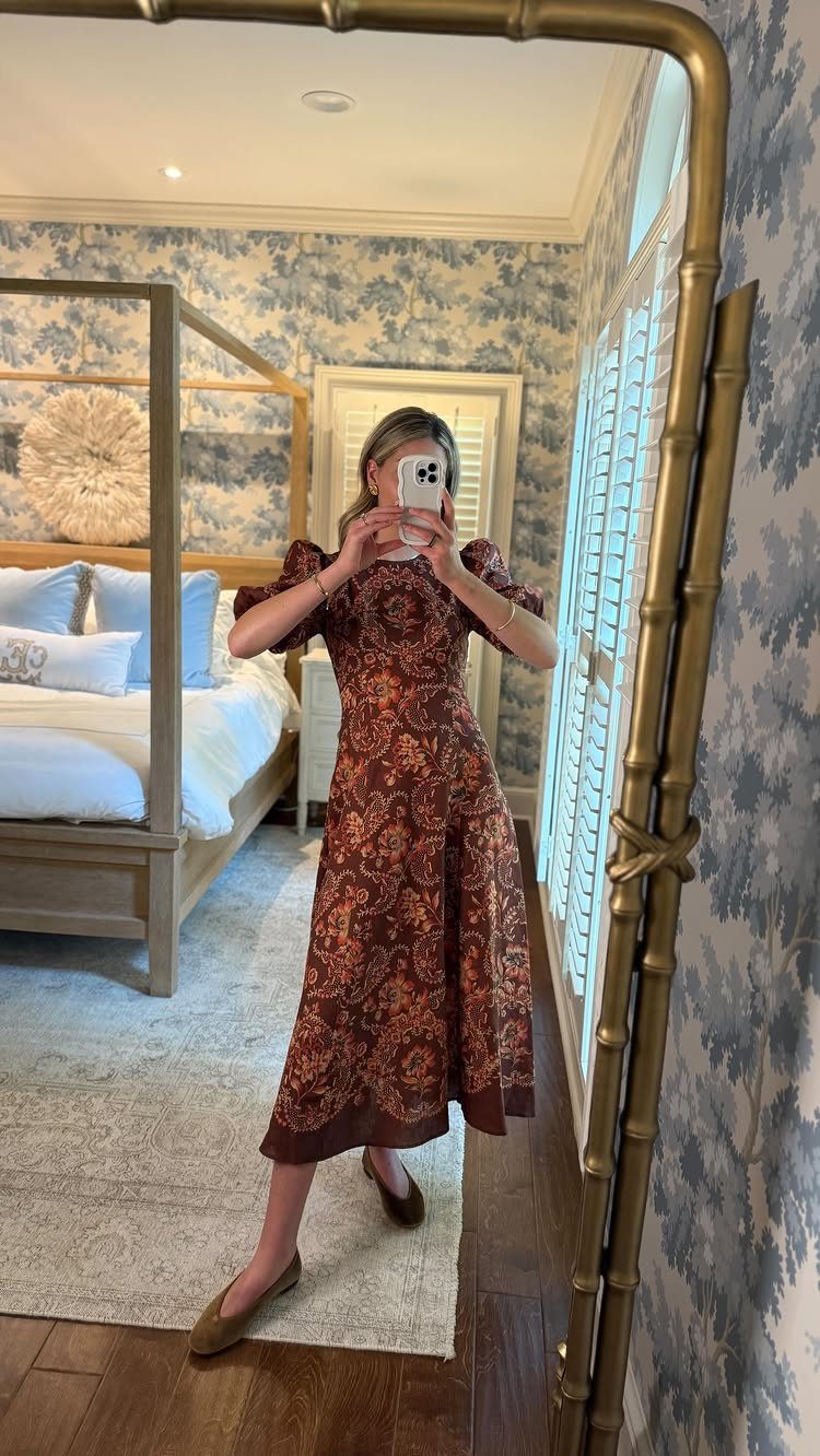 A woman in a patterned reddish-brown midi dress posing for a mirror selfie in a bedroom.