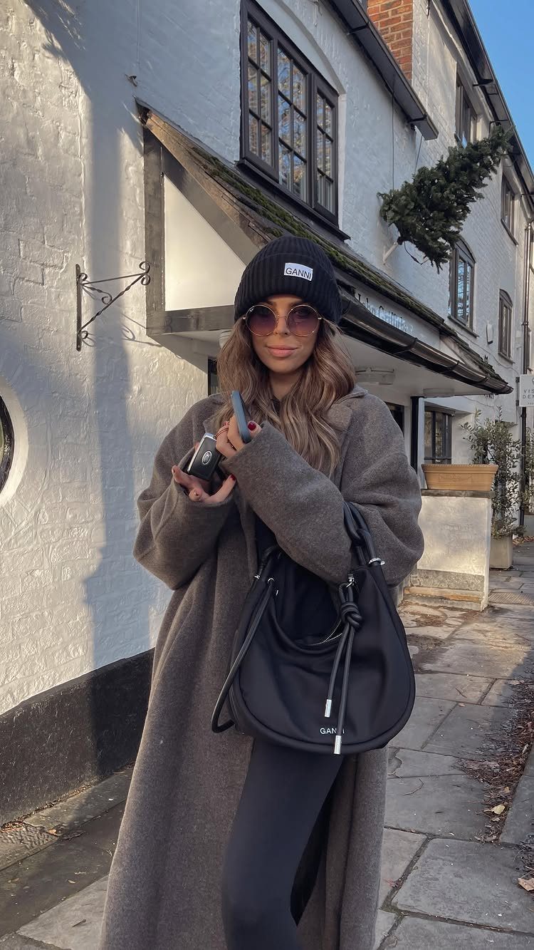 A woman wearing a black beanie, sunglasses, and a long grey coat standing outdoors.