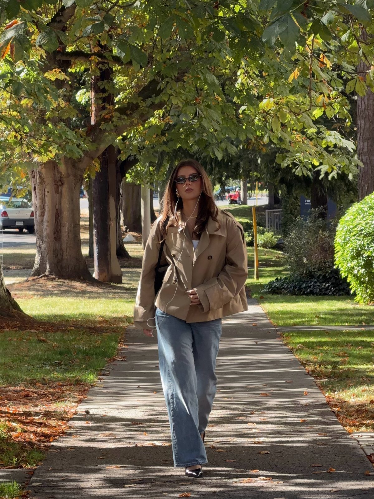 A woman wearing a tan utility jacket and blue jeans walking on a sunny sidewalk.
