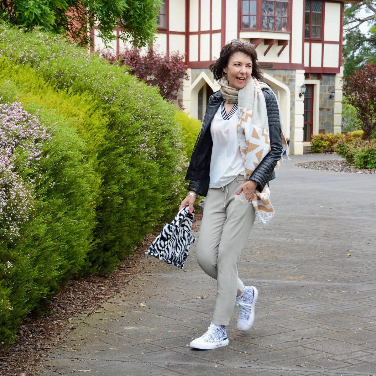 A woman walking outdoors wearing a black leather jacket, white top, and grey trousers.
