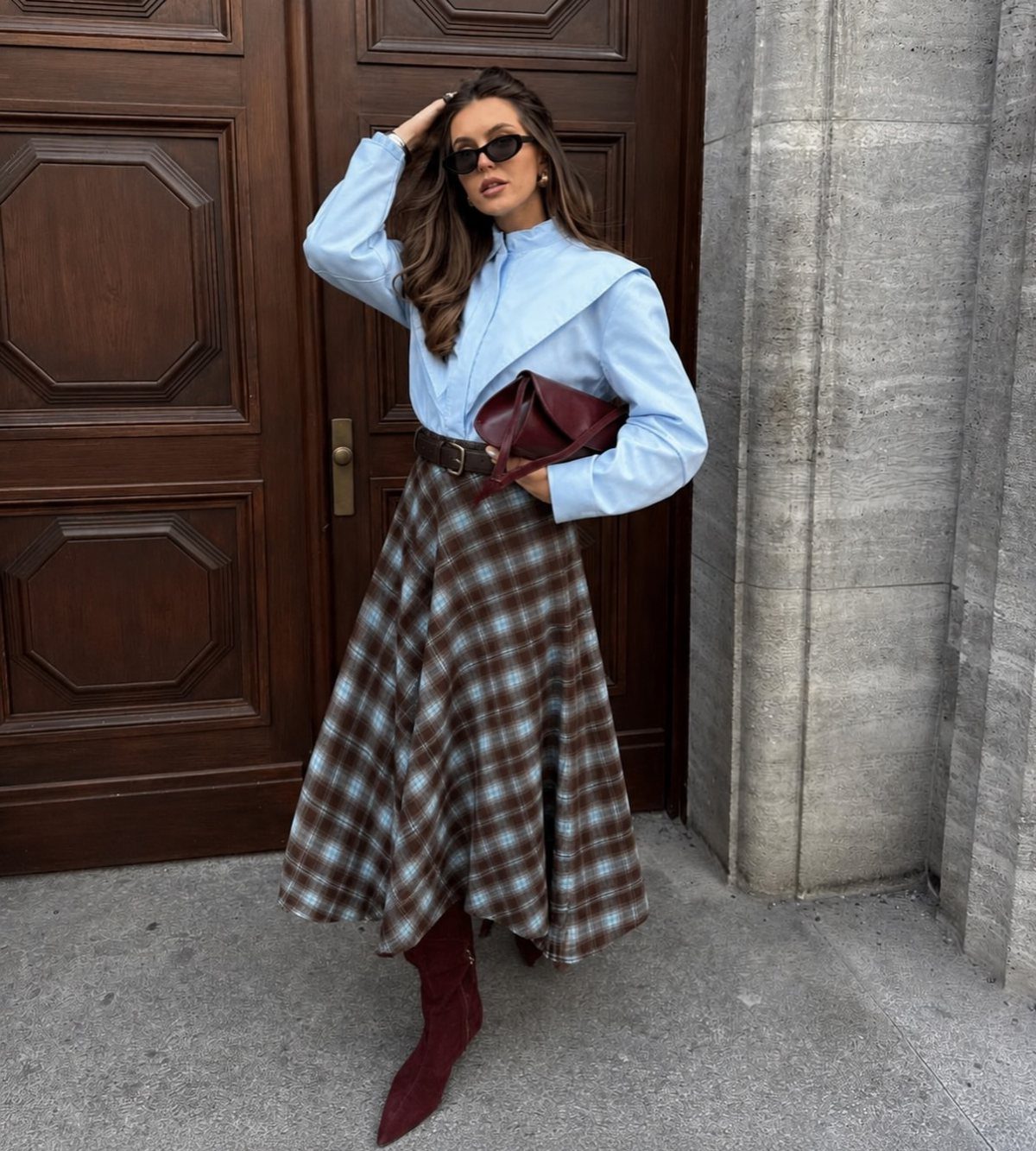 Woman wearing a light blue shirt, plaid midi skirt, and burgundy boots standing by a door.