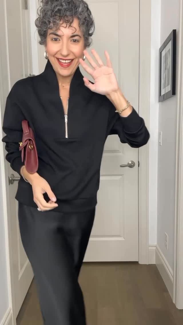Smiling woman with curly hair wearing a black quarter-zip sweater and a black satin slip skirt.