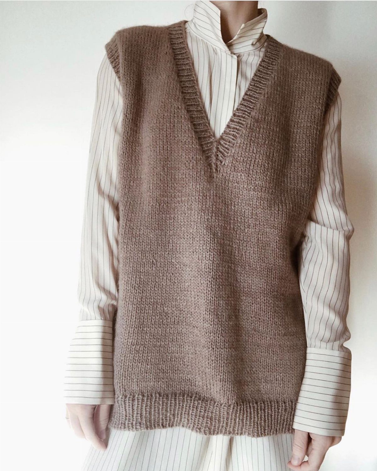 A person wearing a brown knitted sweater vest over a striped long sleeve shirt.