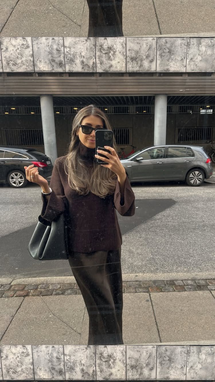 Woman in a dark brown turtleneck sweater and long skirt taking a mirror selfie.