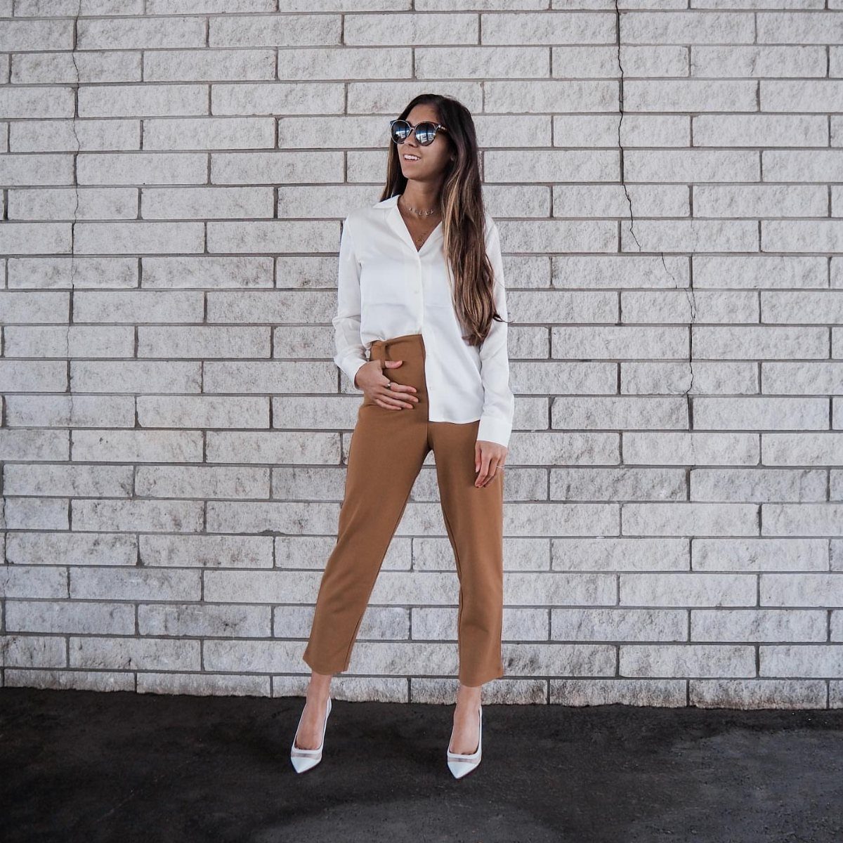A woman in a white blouse and tan trousers posing against a white brick wall.