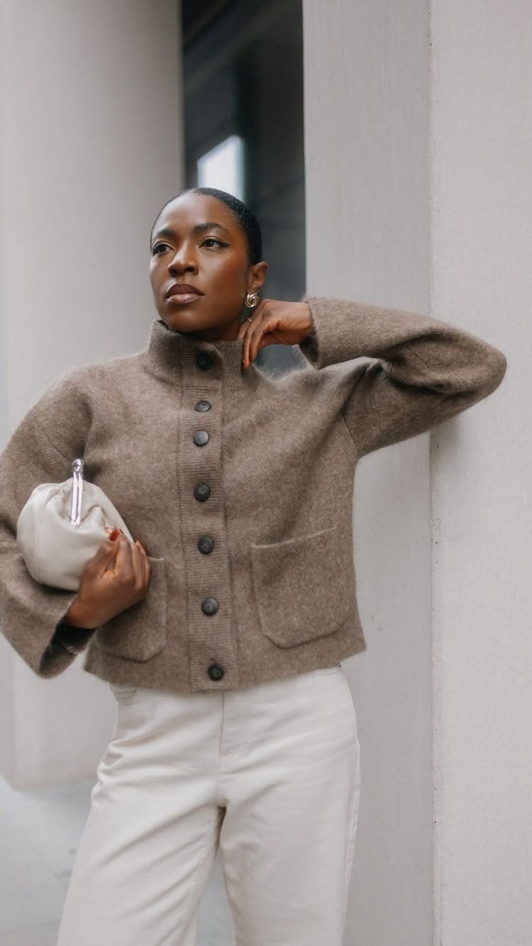 A woman wearing a brown knit button-up jacket and cream trousers standing against a wall.