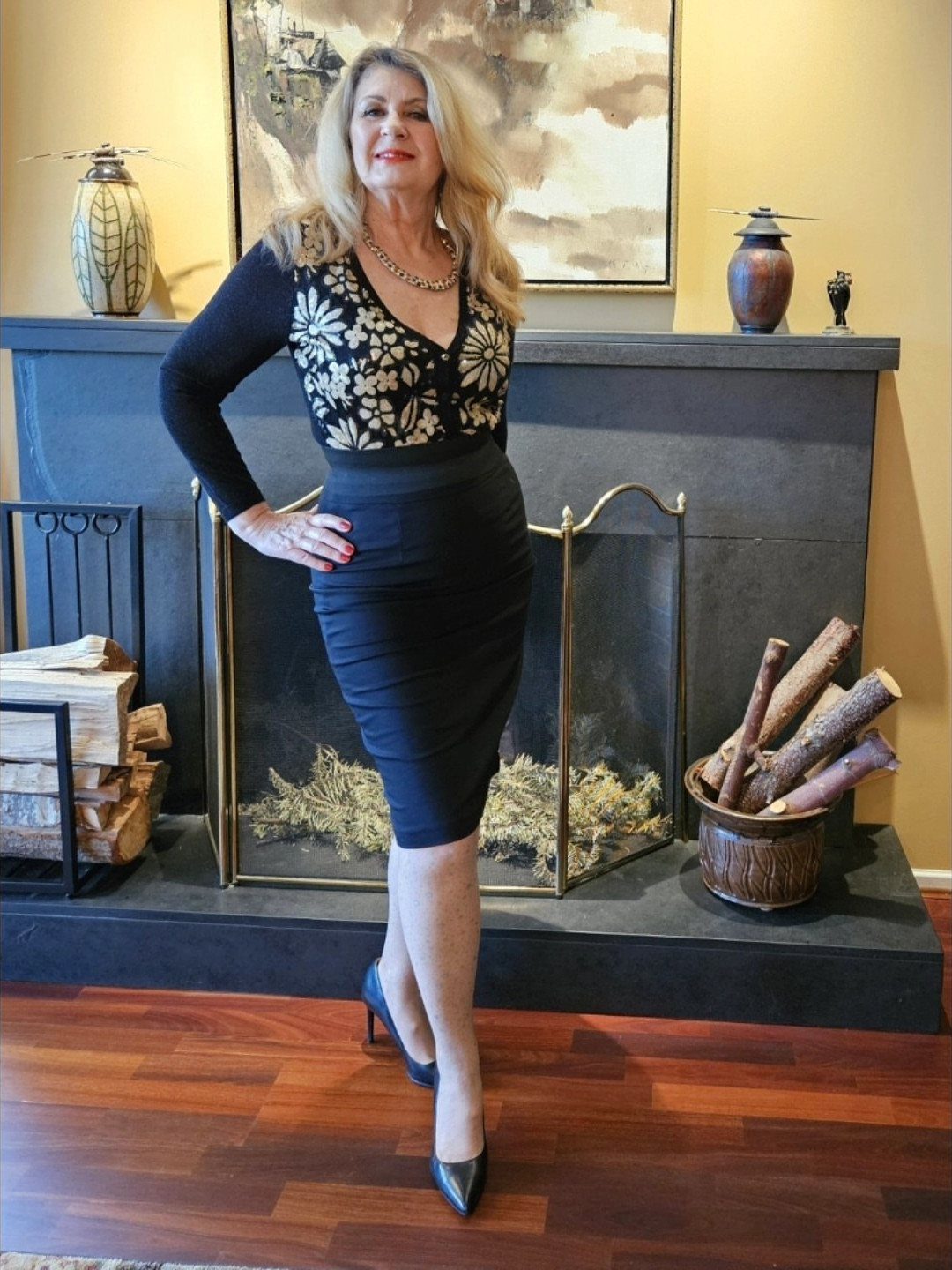 A blonde woman posing in a floral top and black pencil skirt by a fireplace.