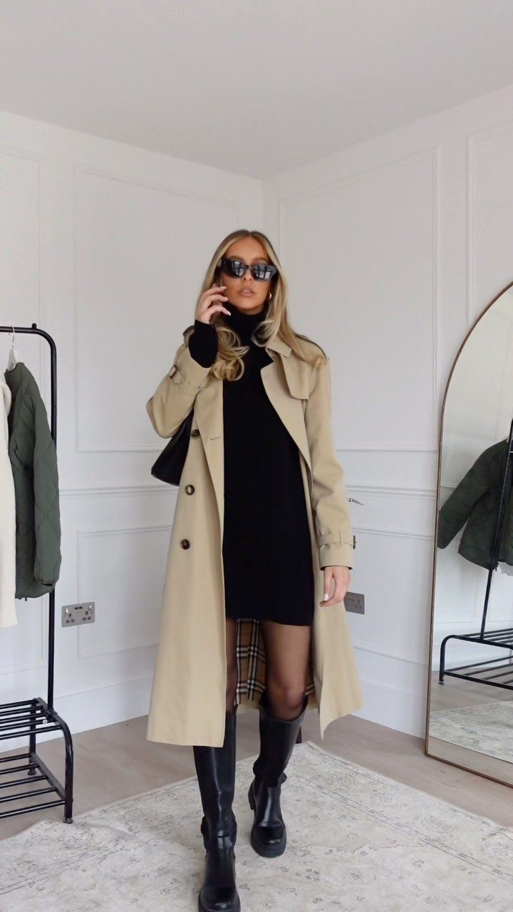 Woman wearing a black turtleneck dress, beige trench coat, and black boots standing indoors.