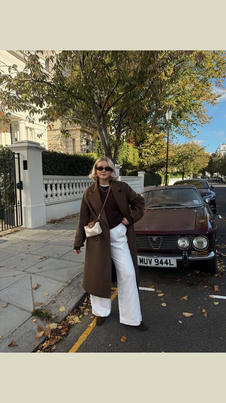 A woman wearing a long brown coat and white wide-leg trousers standing on a city street.