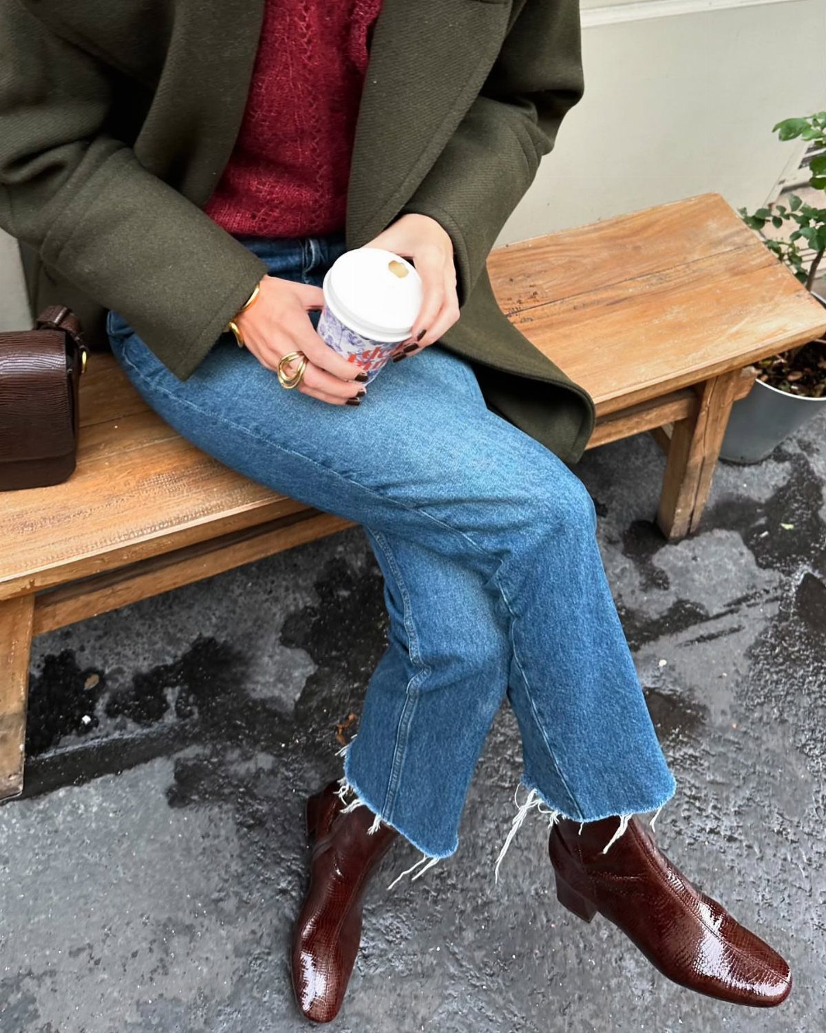 Person wearing blue jeans and dark reddish-brown patent leather ankle boots sitting on a wooden bench.