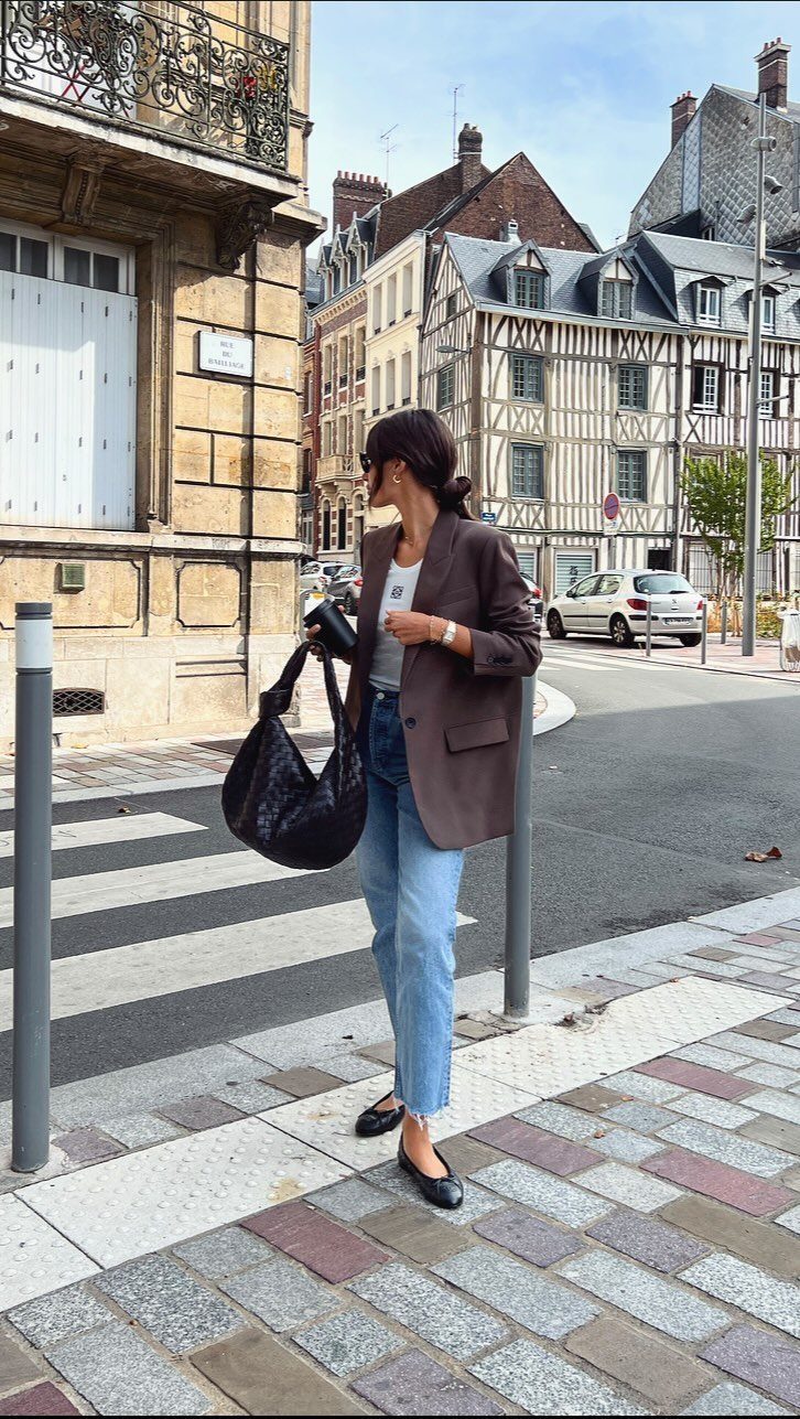 A woman wearing a brown blazer, light blue jeans, and black ballet flats on a city street.